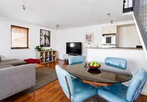 A modern living room with a grey sofa, a glass dining table with blue chairs, a flat-screen TV, and a kitchen area with a white cabinet and a small plant.
