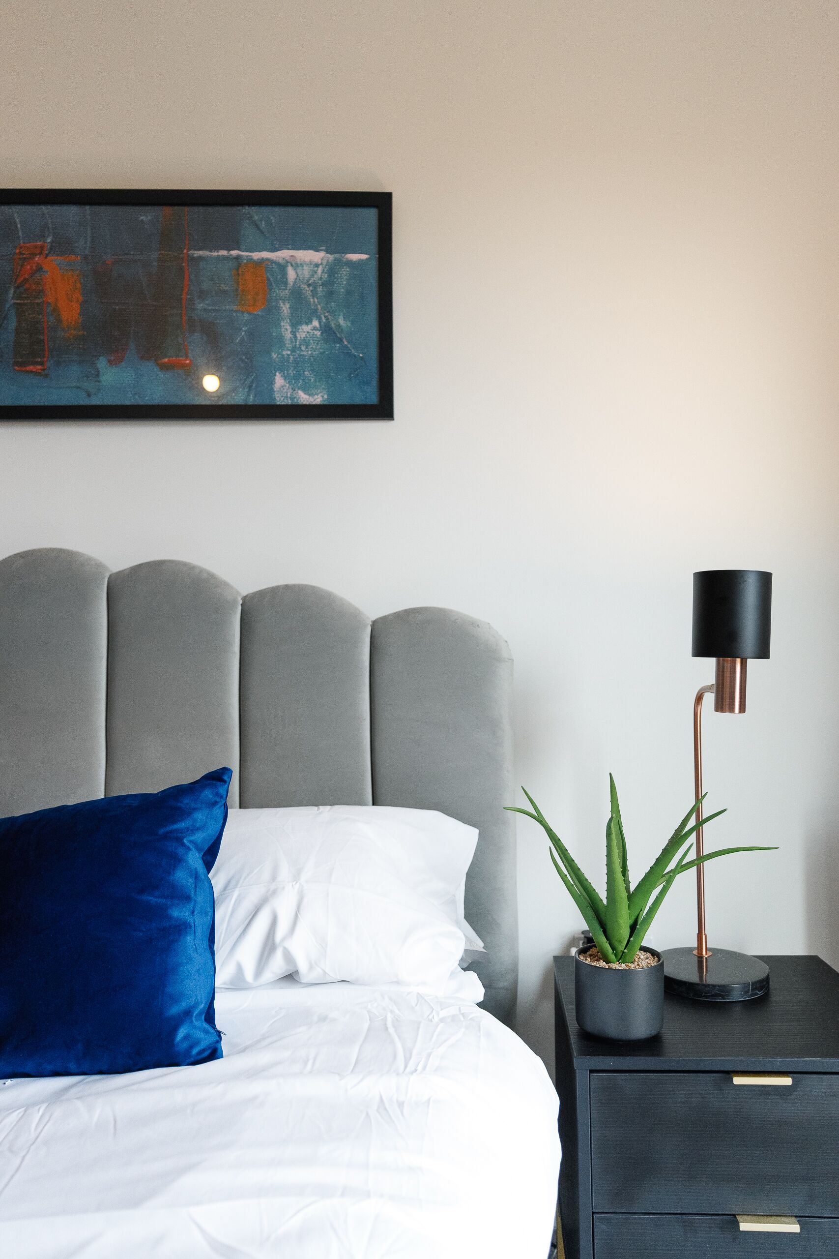 A neatly made bed with a blue pillow and white sheets, a grey upholstered headboard, a black lamp on a side table, and a potted plant.