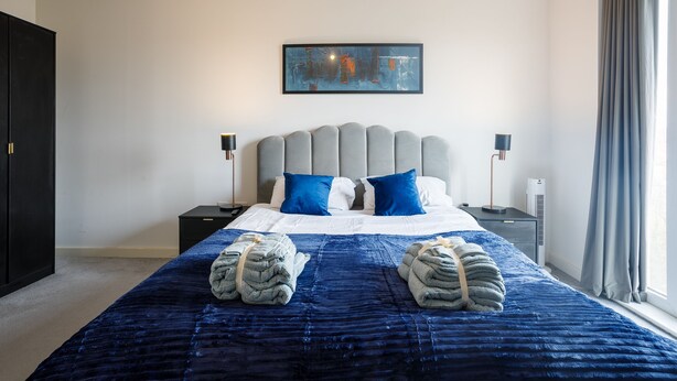 A modern bedroom with a large bed, grey upholstered headboard, blue and white bedding, and a painting above the bed. - Charming 1-bed Apartment in London (London)