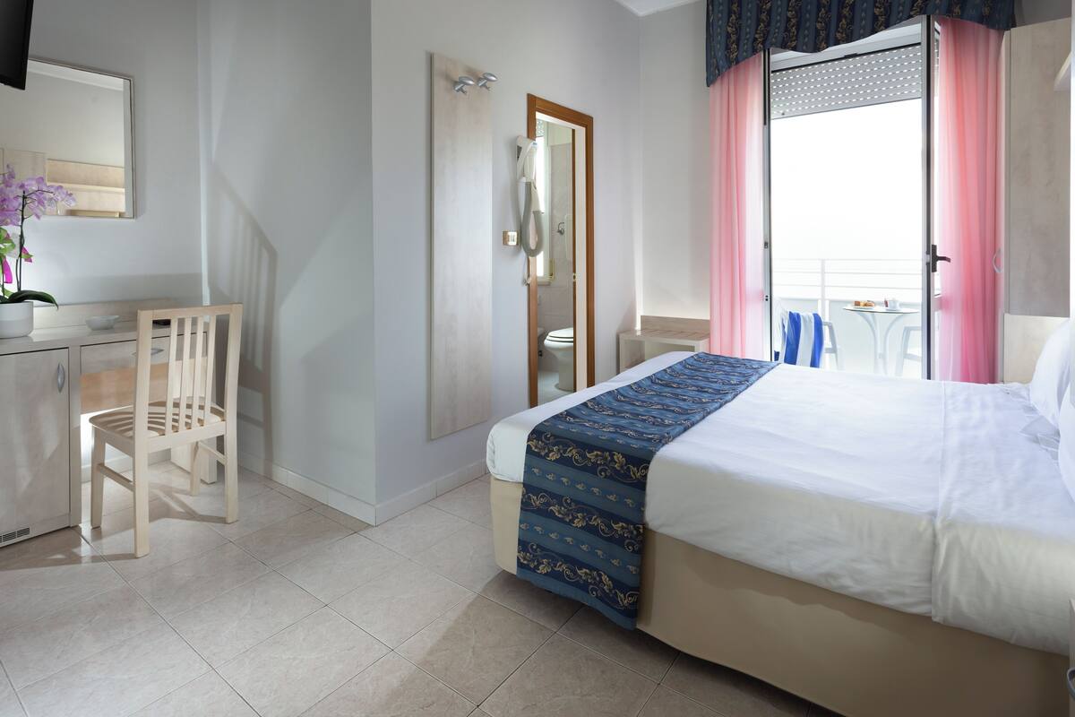 A hotel room with a bed, a desk, a chair, a bathroom, and a balcony with a view.