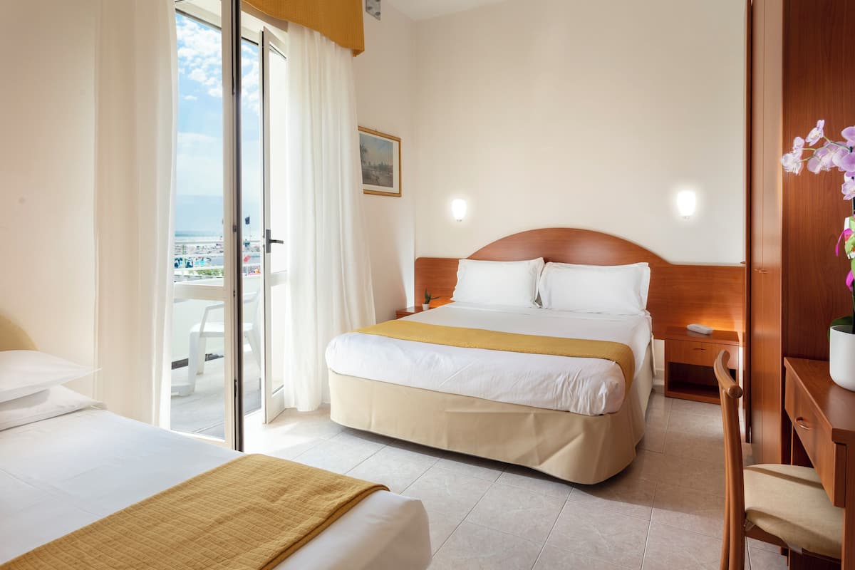 A hotel room with two beds, a wooden headboard, a nightstand, a desk, and a view of the beach through a sliding door.