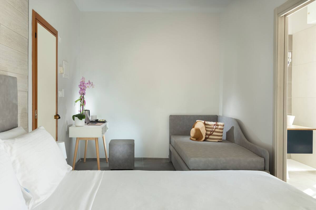 A modern hotel room with a bed, a desk, a grey sofa, and a small stool.
