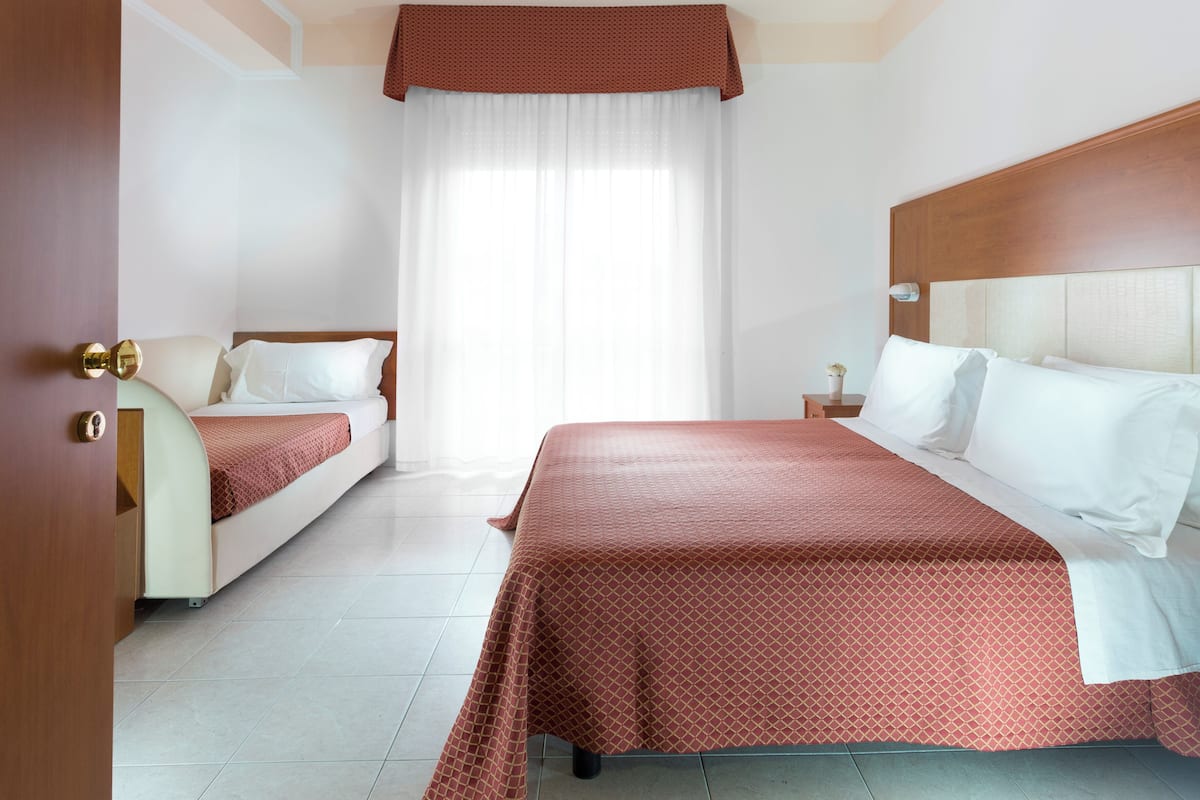A hotel room with two beds, a large window with sheer curtains, a wooden headboard, and a bedside table with a lamp.