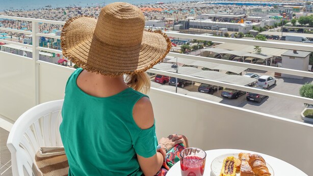 A person seated at a table with a view of a beach and cityscape. - Hotel Vela d'Oro (Rimini)