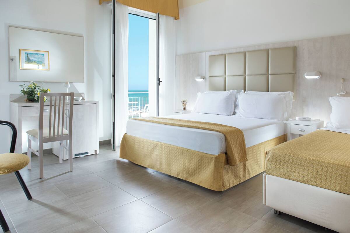A hotel room with a bed, a desk, a chair, and a view of the sea.