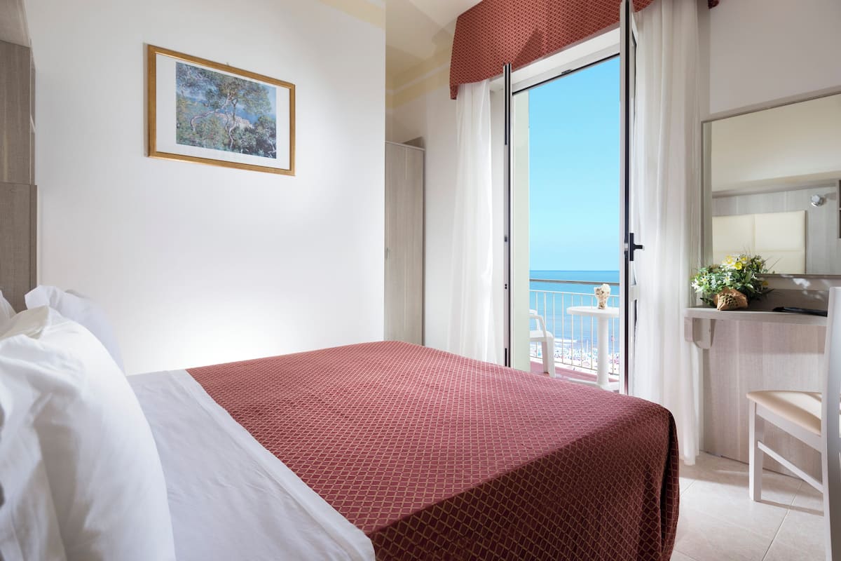A hotel room with a bed, a small desk, a chair, a balcony with a view of the sea, and a framed picture on the wall.