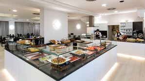 A buffet area with various food options, a seating area with tables and chairs, and a kitchen in the background. - Hotel Vela d'Oro (Rimini)