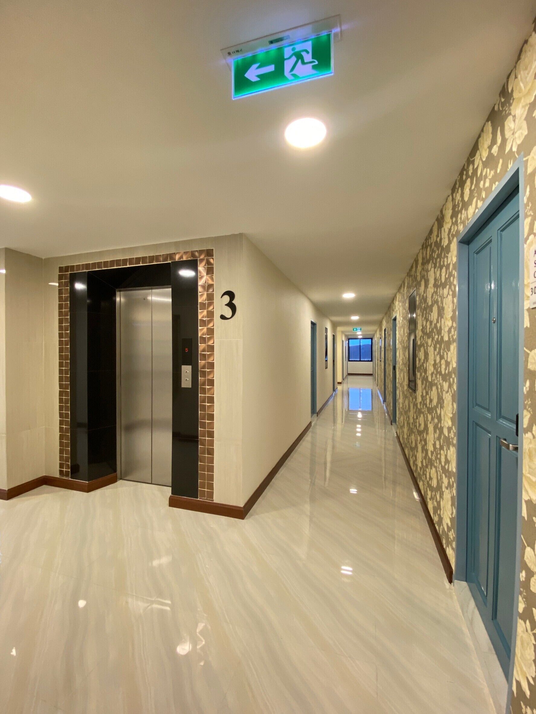 Corredor de hotel com piso lustroso, placa com o número 3, um elevador e portas azuis.