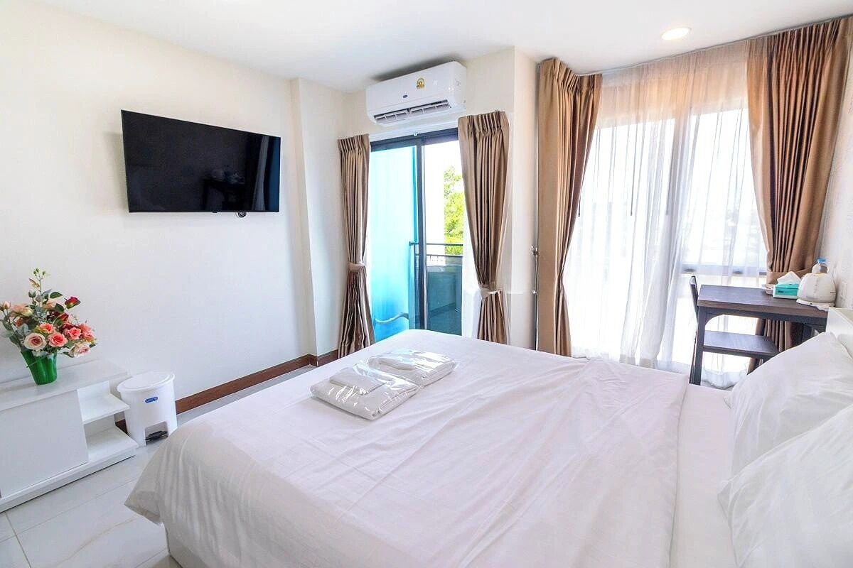 A hotel room with a bed, a flat-screen TV mounted on the wall, an air conditioning unit, a window with curtains, a small table with a chair, and a vase with flowers.