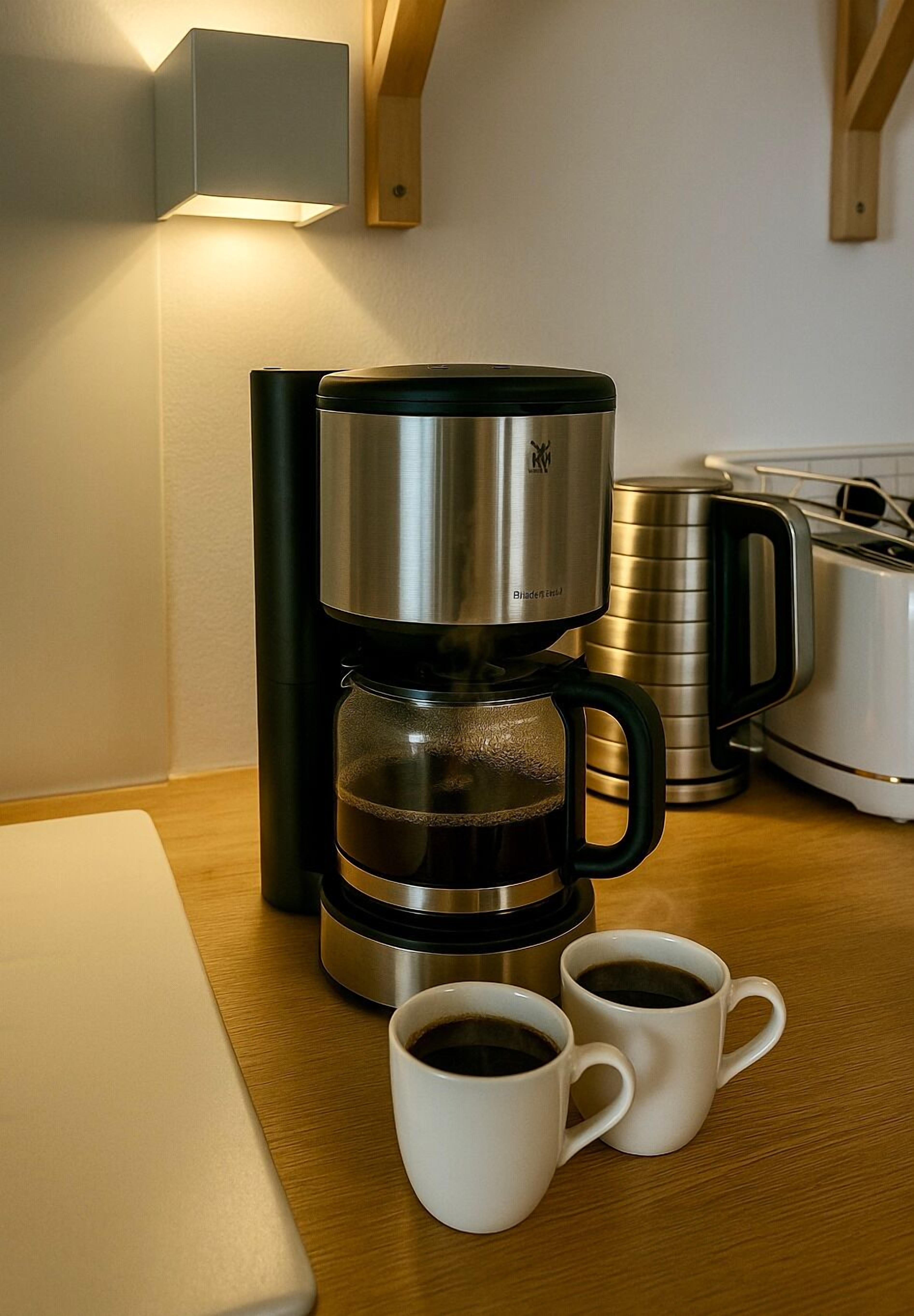 Máquina de café com duas chávenas brancas sobre uma mesa de madeira, torradeira e chaleira numa cozinha.