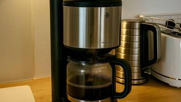 Máquina de café com duas chávenas brancas sobre uma mesa de madeira, torradeira e chaleira numa cozinha.