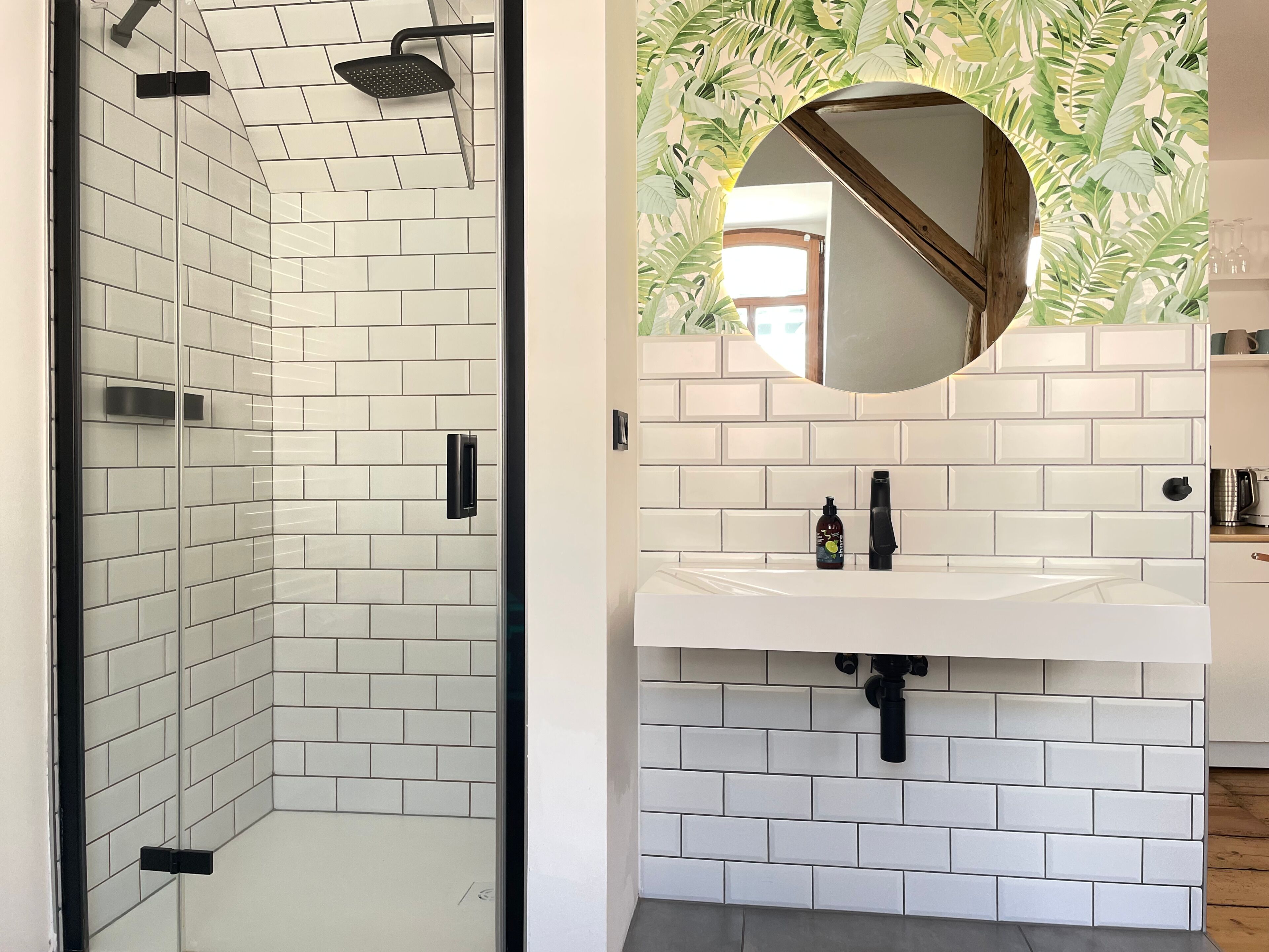 A modern bathroom with white subway tiles, a glass-enclosed shower, a round mirror, and a sink with a wooden countertop.