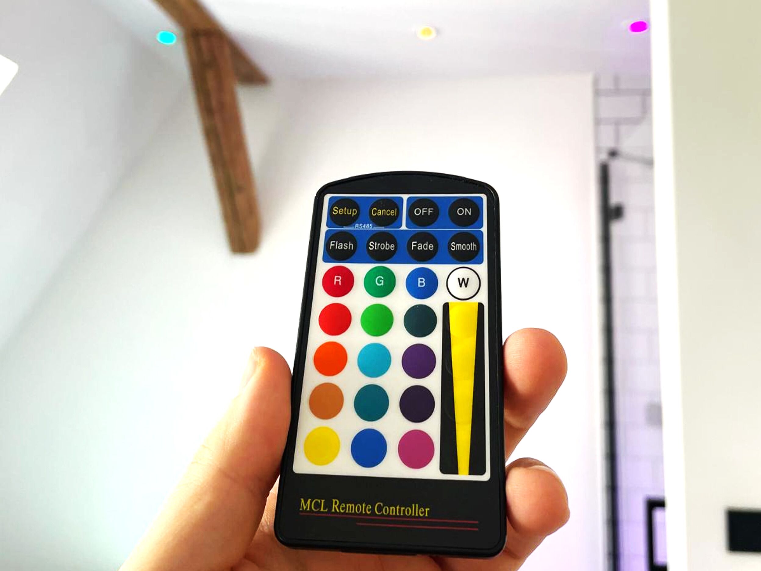 En hånd holder en fjernbetjening med flere farveknapper og en etiket der står 'MCL Remote Controller'.