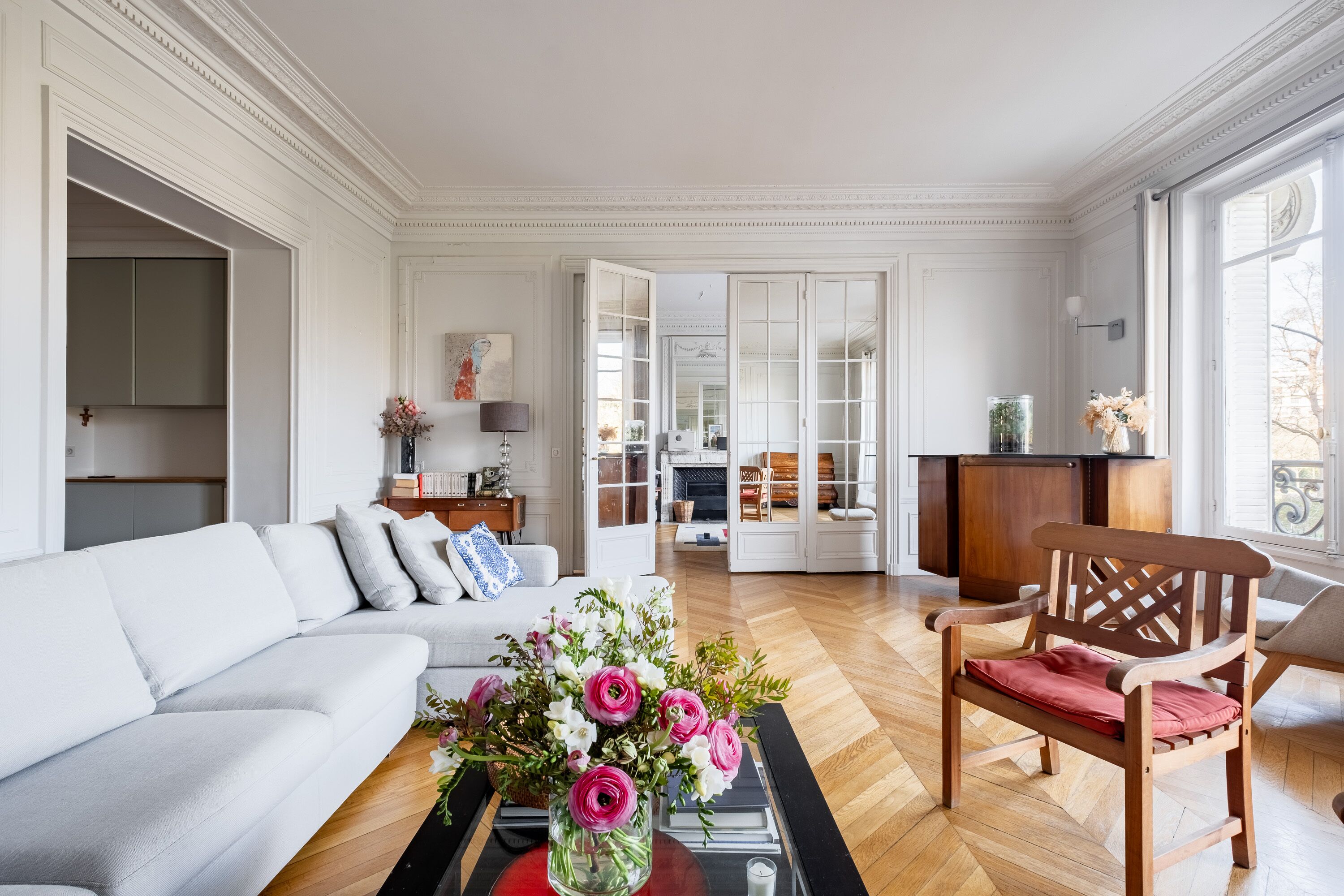 A spacious living room with a light grey sofa, a wooden chair, and a coffee table with a vase of flowers.