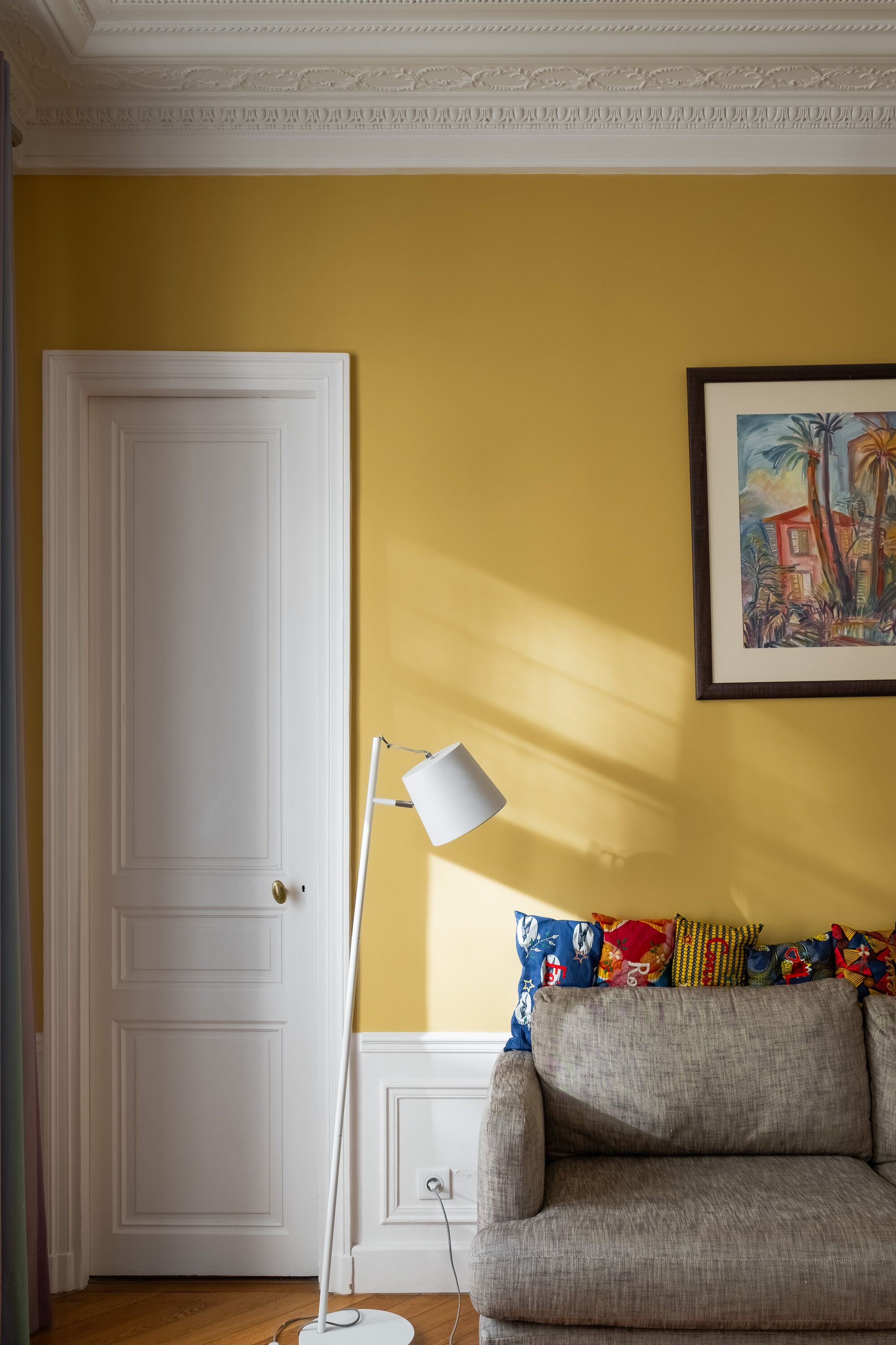 A room with a yellow wall, a white door, a gray sofa with colorful pillows, a white floor lamp, and a framed painting.
