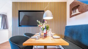 A dining area with a wooden table, two chairs, a vase with flowers, and a television mounted on the wall. - Watt. Familie. Reet (Keitum)