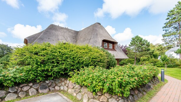 A house with a steeply pitched roof, surrounded by dense greenery and a stone wall. - Watt. Familie. Reet (Keitum)