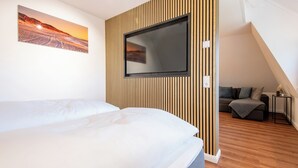 A modern hotel room with a large bed, a flat-screen TV mounted on a wooden panel wall, a painting of a sunset, and a cozy seating area with a sofa and a small table. - Watt. Familie. Reet (Keitum)