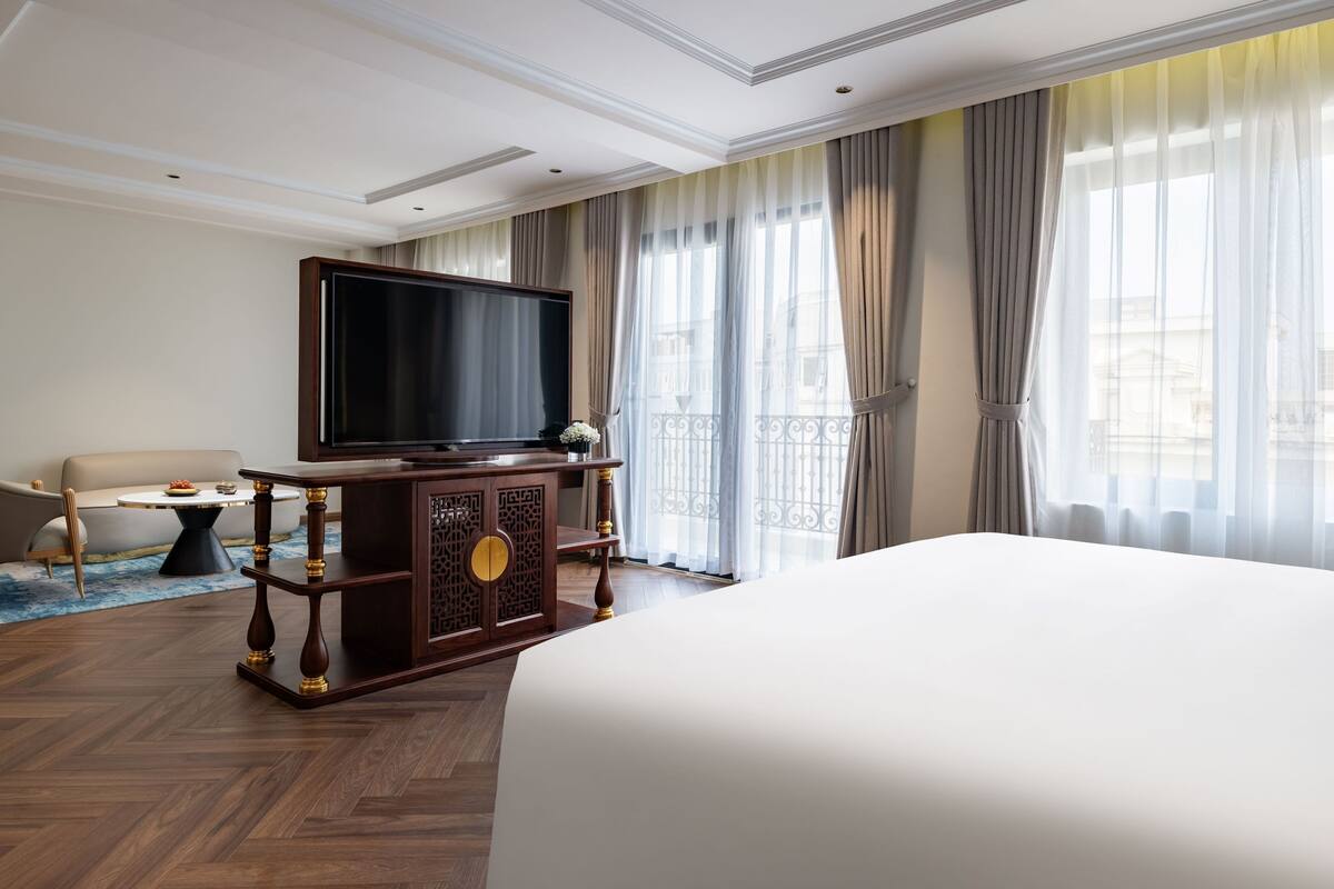 A hotel room with a large bed, a television on a wooden console, a sofa, and a view of the city through sheer curtains.