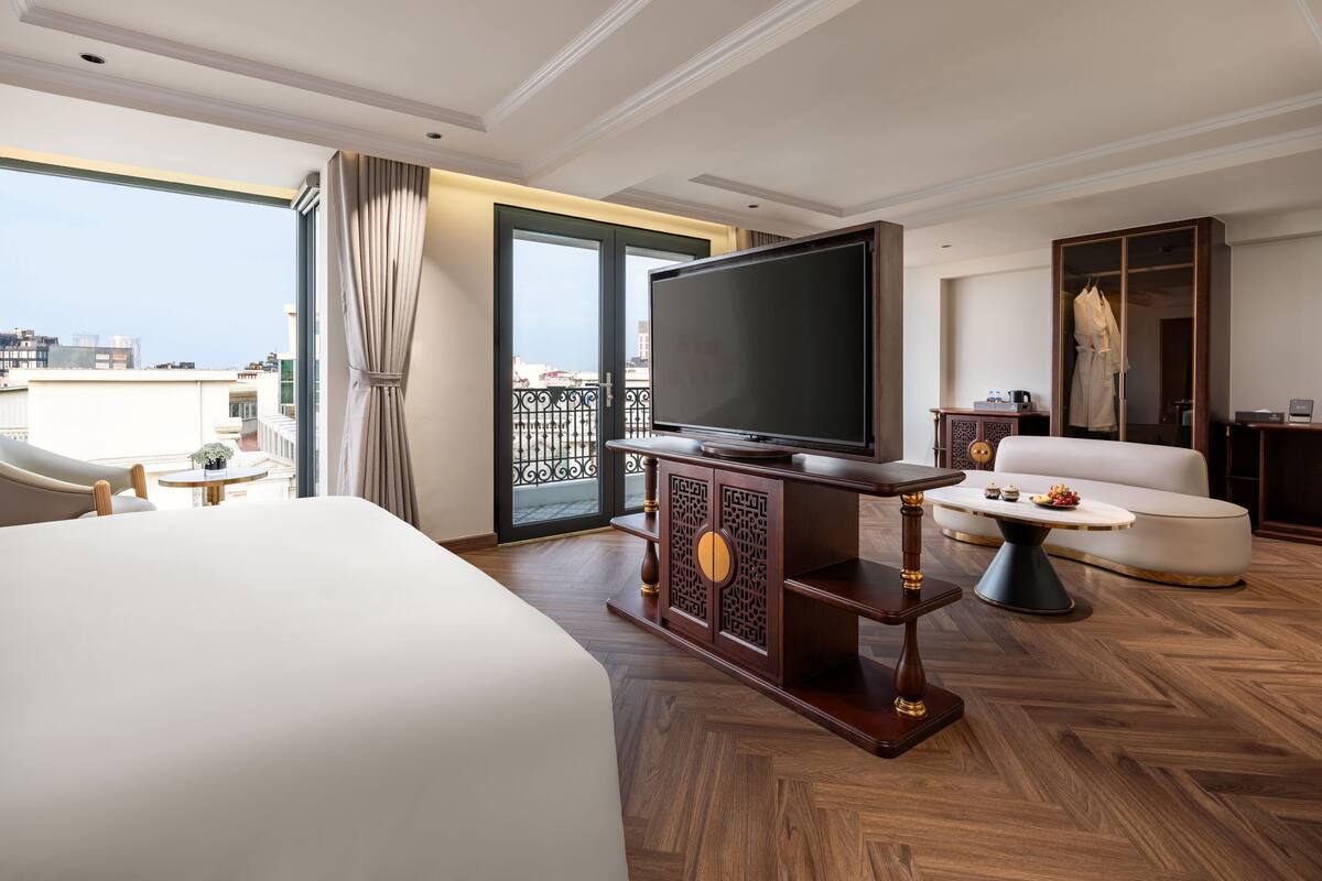 A hotel room with a large bed, a television on a wooden stand, a sofa, and a view of the city through a balcony.