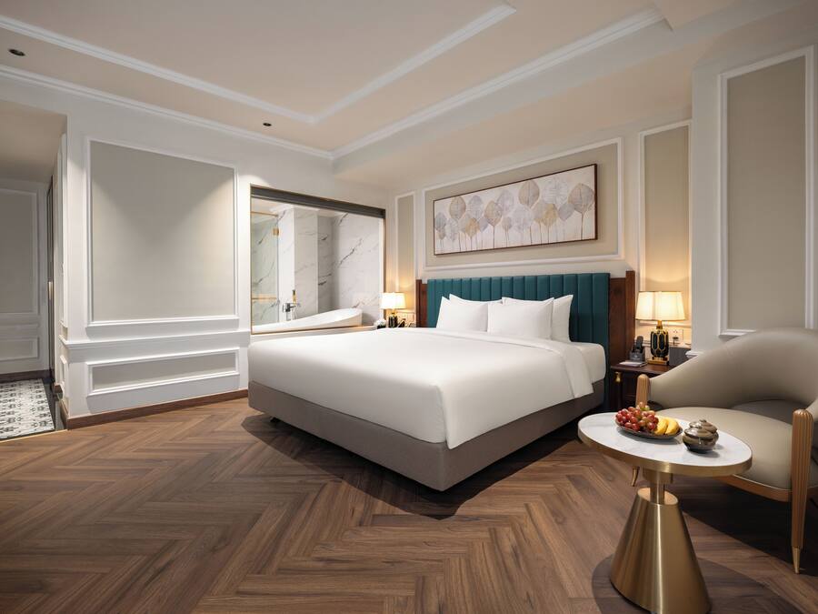 Habitación de hotel con una cama grande, una zona de estar con una silla, una pequeña mesa con fruta y un baño con bañera y espejo.