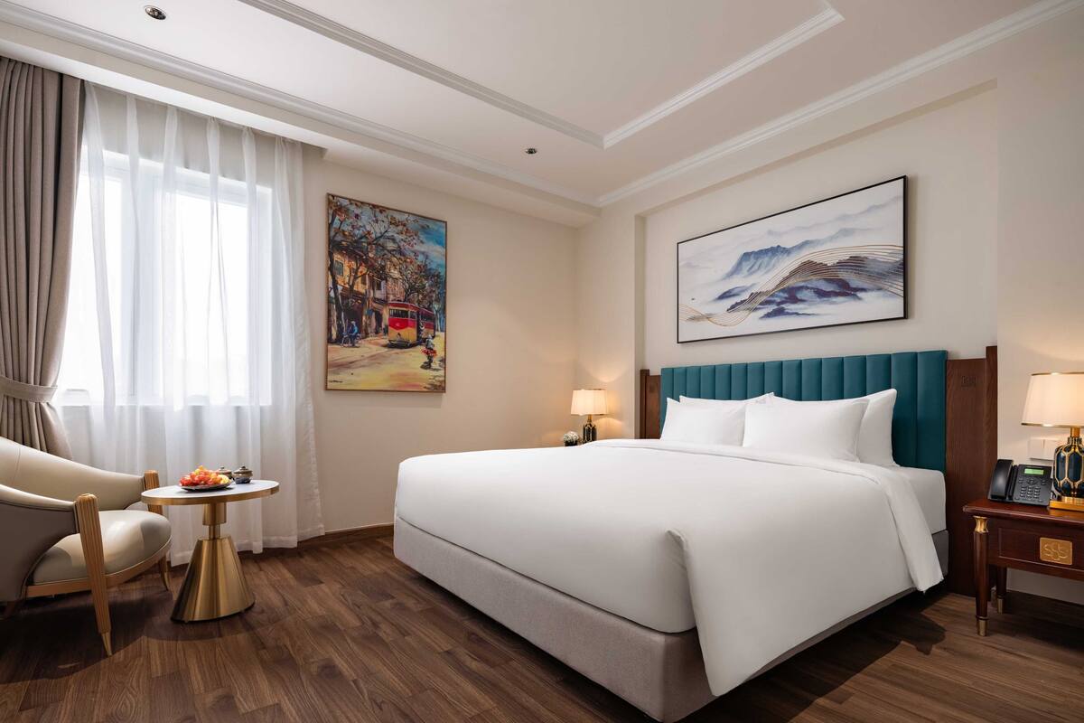 A hotel room with a large bed, a chair, a small table with fruit, and artwork on the walls.