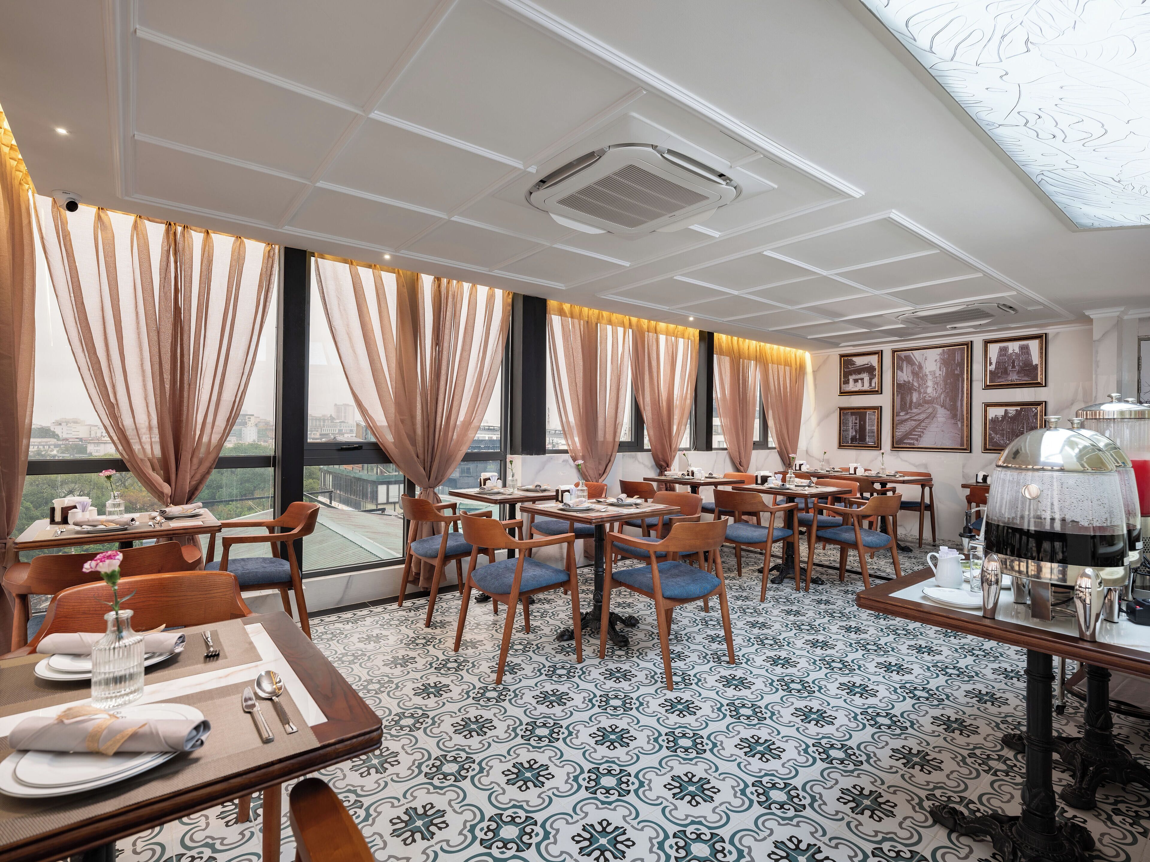 A restaurant with tables set for dining, large windows with curtains, and a patterned floor.