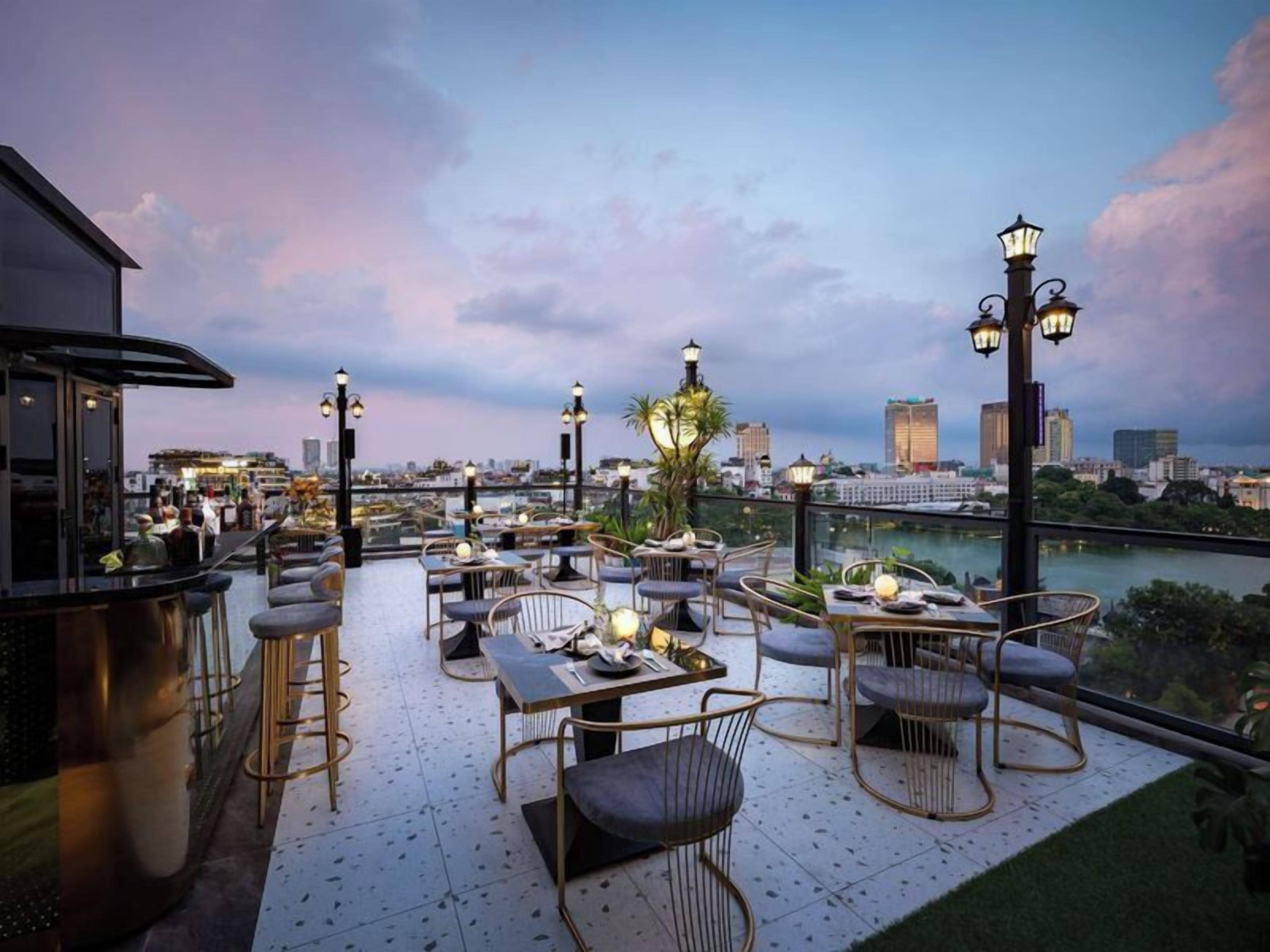 An outdoor dining area with tables and chairs, a view of the city skyline, and a river in the background.
