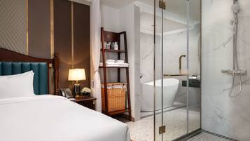 A hotel room with a bed, bedside table, lamp, and a glass-enclosed shower area with a bathtub.