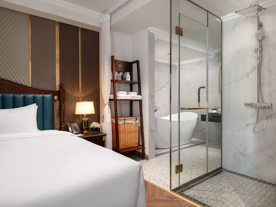 A hotel room with a bed, bedside table, lamp, and a glass-enclosed shower area with a bathtub.