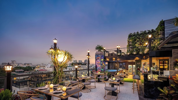 Una terraza en la azotea con mesas y sillas, un mural y vistas de la ciudad al anochecer.