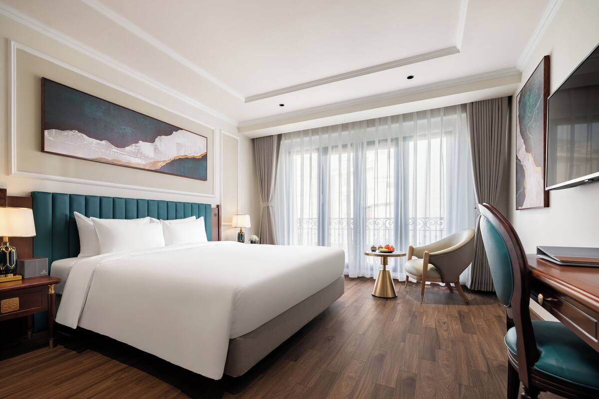 A hotel room with a large bed, a desk, a chair, a small table with a bowl of fruit, and a large window with curtains.