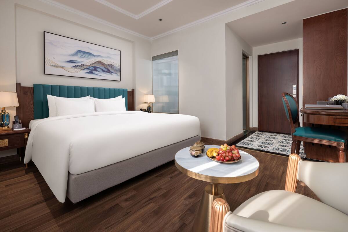 A hotel room with a large bed, a round coffee table with fruit, a desk with a chair, and a painting on the wall.