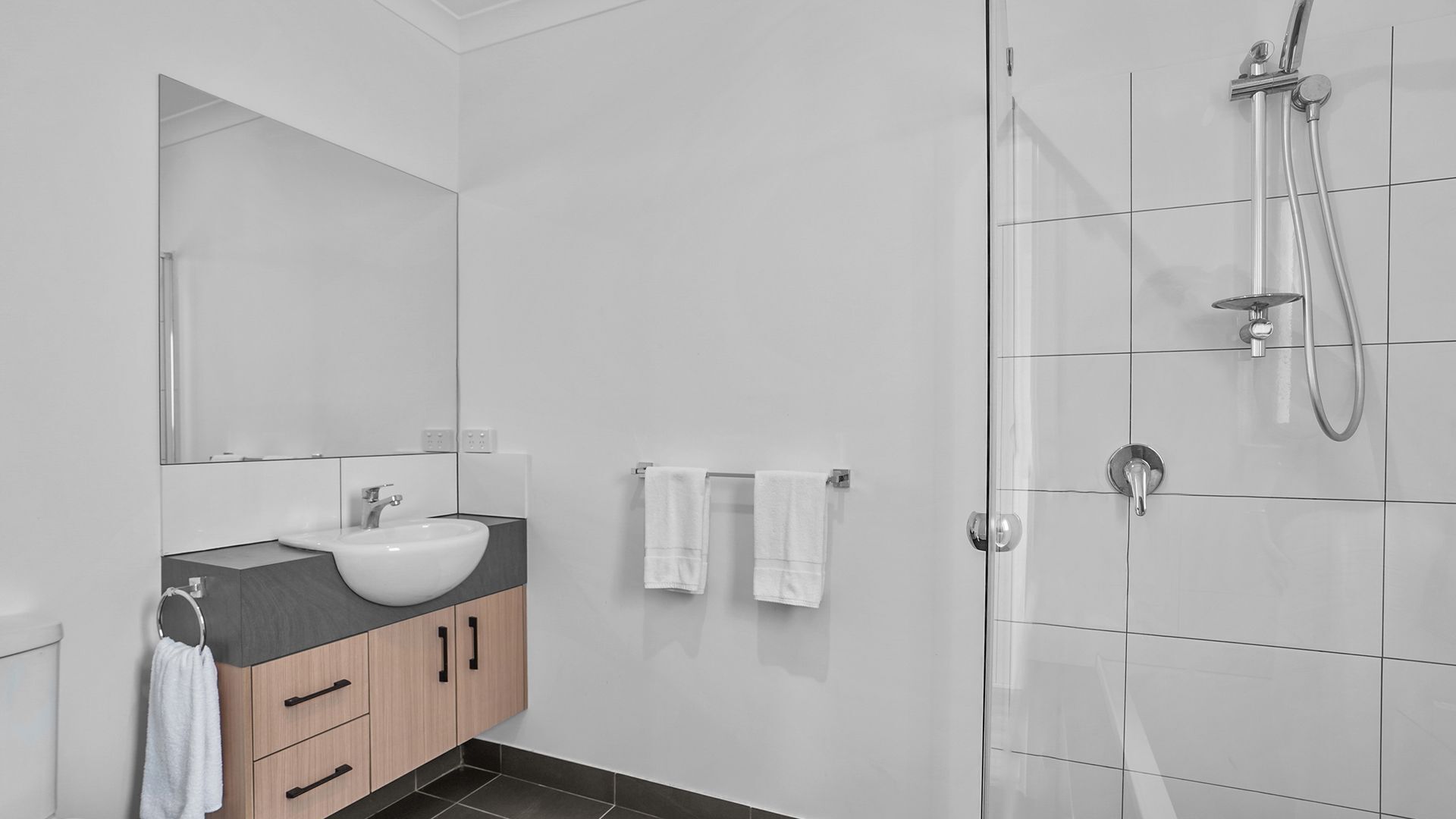 Un baño moderno con un lavabo blanco, armarios de madera, un espejo grande y una ducha a ras de suelo.