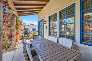 Eine überdachte Terrasse mit einem Holztisch und Korbstühlen, umgeben von blauen Fensterrahmen und einem Hängesessel.