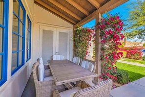 Eine überdachte Terrasse mit Holztisch und Korbstühlen, eine weiße Tür mit Fensterläden und Blick auf einen Garten mit rosa Blumen.