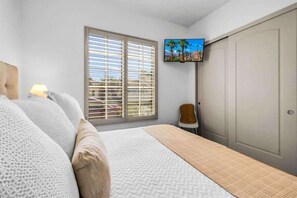 Ein Schlafzimmer mit einem Bett, einem an der Wand montierten Fernseher, einem Fenster mit Jalousien und einem Kleiderschrank.