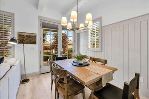 Comedor con mesa de madera, sillas, una lámpara de araña y vistas a través de un ventanal.