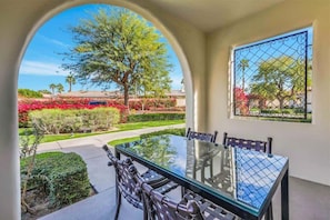 A dining area with a glass table and chairs, an arched doorway leading to a garden with a tree and red flowers. - Modern Palms B | Studio Legacy Villa (La Quinta)