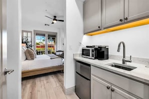 Een kleine keuken met een magnetron, spoelbak en kastjes. Een bed is zichtbaar door een geopende deur, en er hangt een plafondventilator.