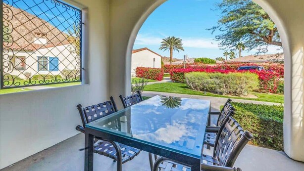 Mesa con cubierta de vidrio y cuatro sillas en un espacio exterior cubierto, con vista a un jardín ajardinado con flores rojas y una palmera.