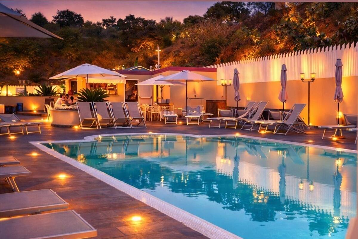 A swimming pool with lounge chairs and umbrellas, surrounded by a landscaped area with trees and a building in the background.
