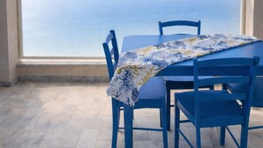 Een tafel met blauwe stoelen en een tafelkleed, voor een groot boogvenster met uitzicht op zee.