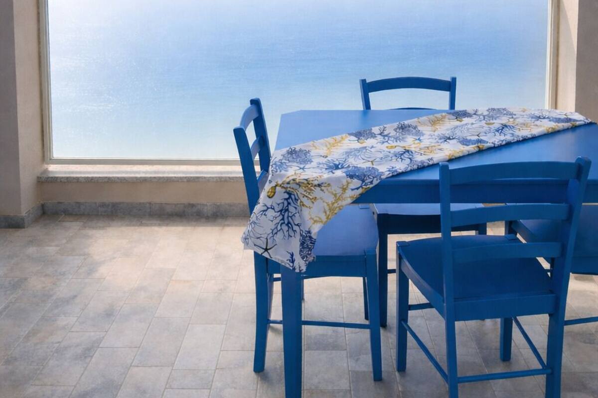 A table with blue chairs and a tablecloth, set against a large arched window overlooking the sea.