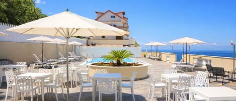 An outdoor patio with white tables and chairs, umbrellas, and a pool area.