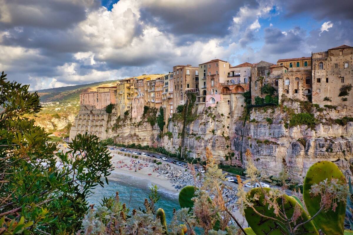 A coastal town with buildings built into the cliffside, overlooking a beach with people and cars.