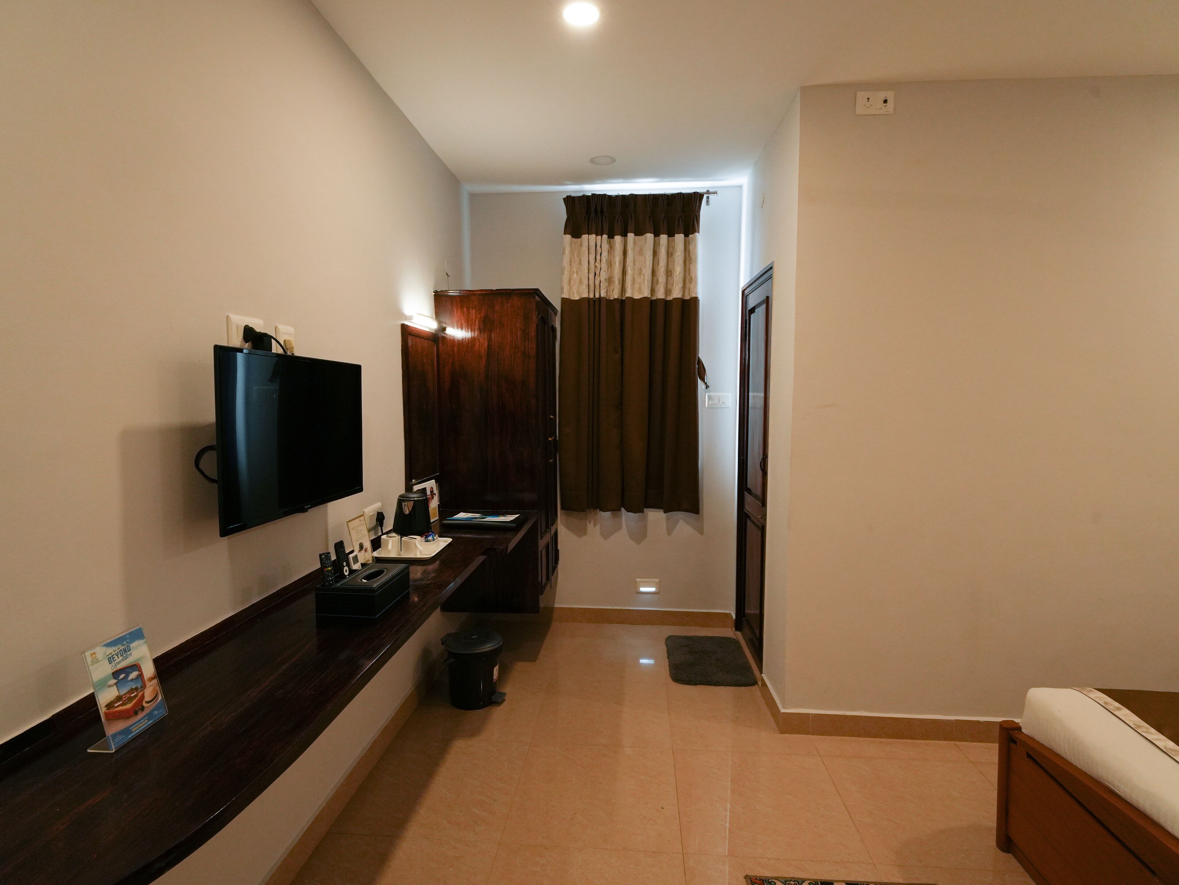 A hotel room with a dark wooden vanity, a flat-screen TV mounted on the wall, a small trash bin, and a bed with a white mattress.