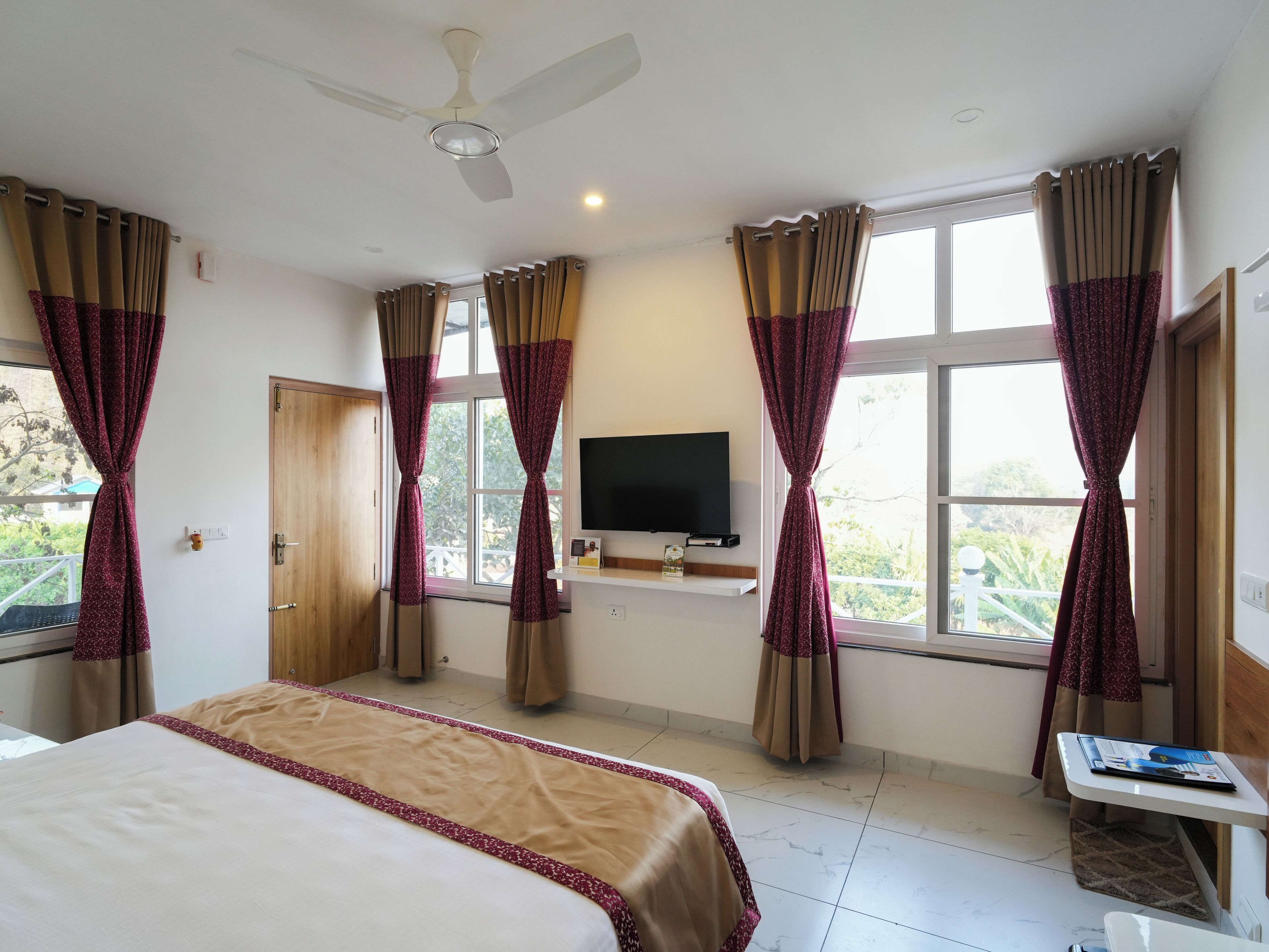 A hotel room with a bed, a ceiling fan, a TV mounted on the wall, and large windows with curtains.