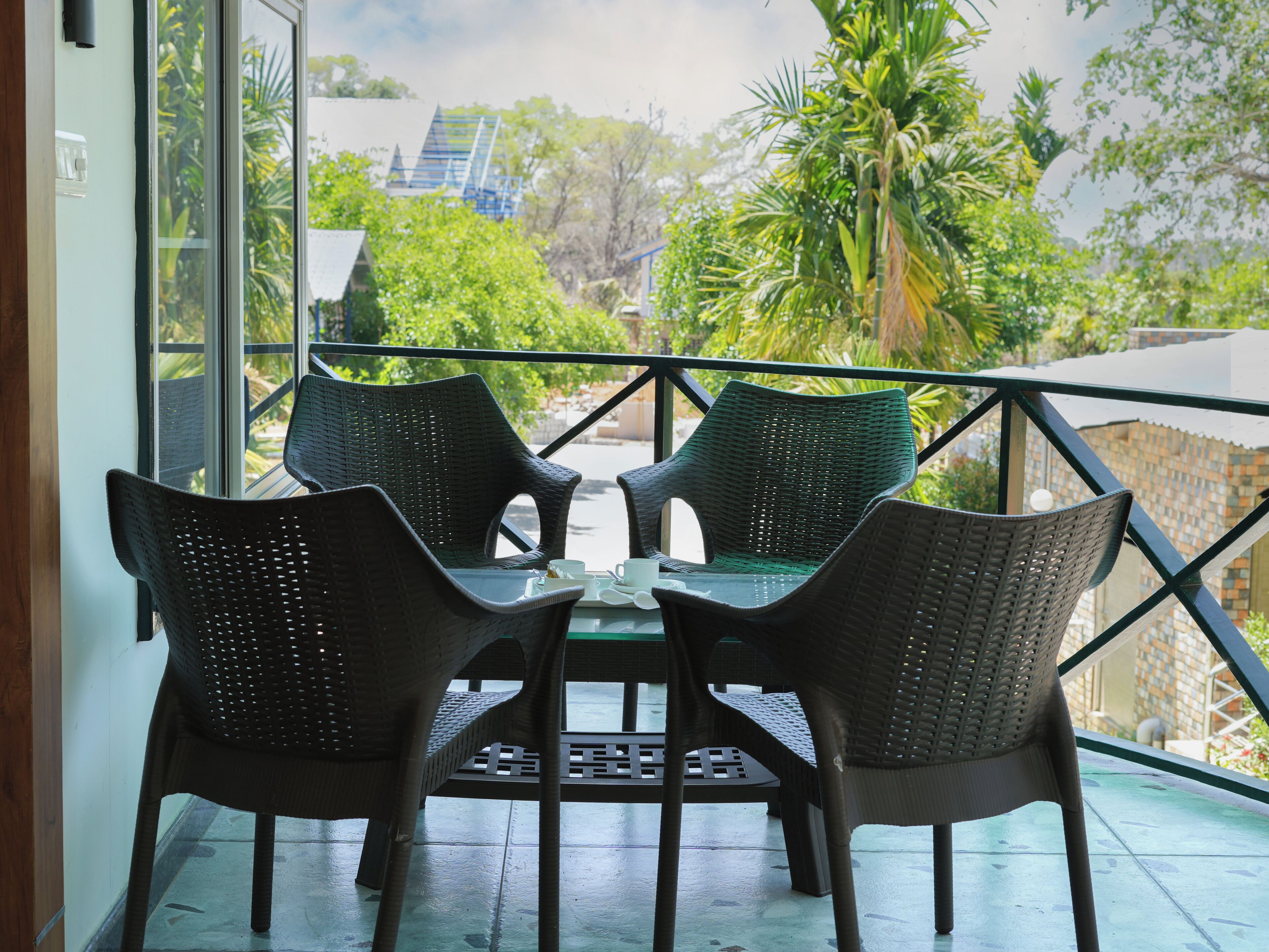 A balcony with four wicker chairs and a small table, overlooking a lush garden and a distant building.