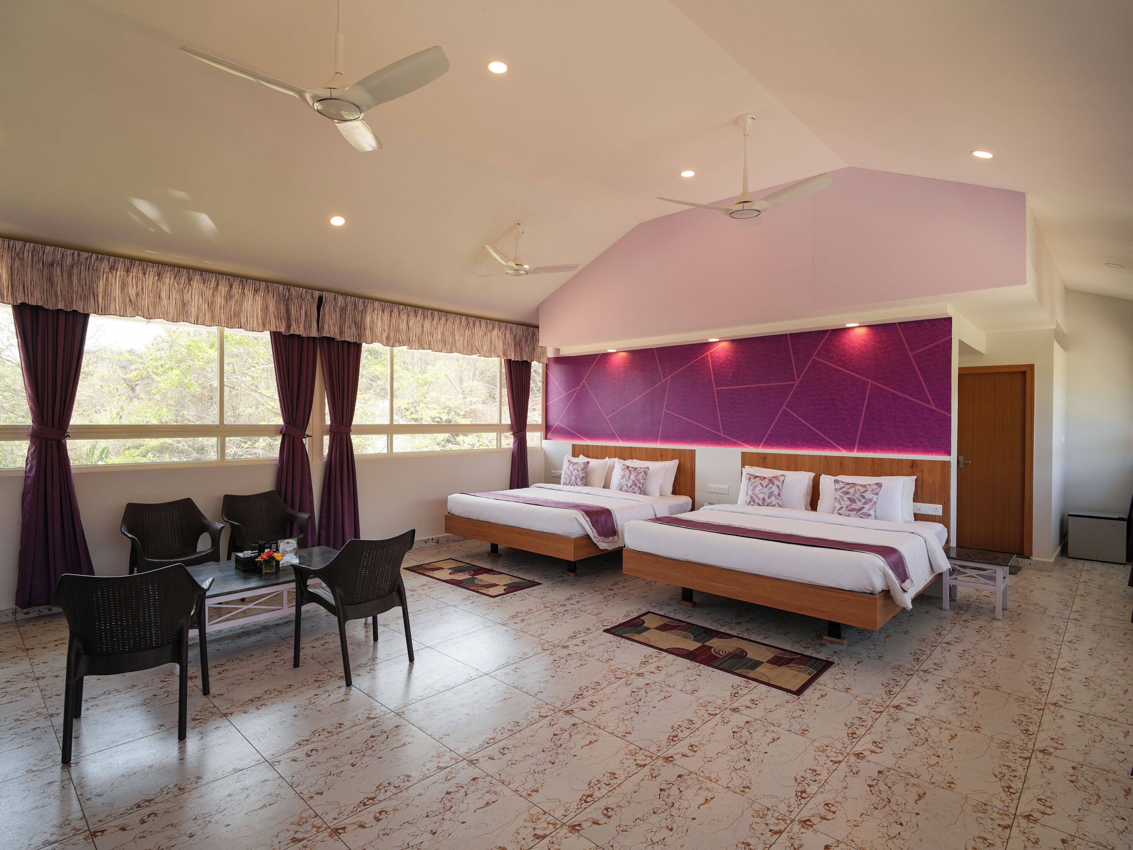 A bedroom with two beds, a dining area with chairs, a ceiling fan, and a view of trees through the window.